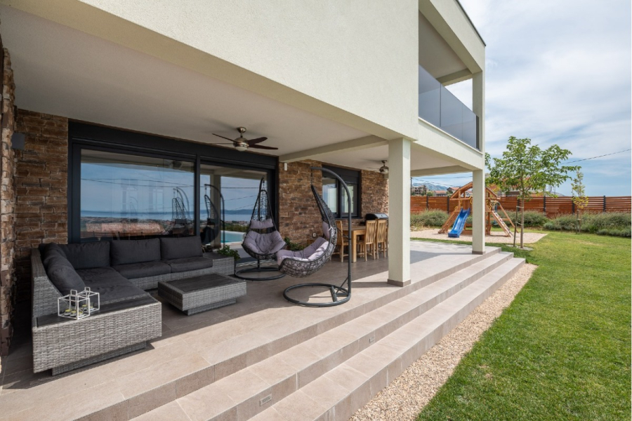 Villa am Stadtrand von Split großzügige Aufteilung, hohe Räume und beheiztem Pool - Moderne überdachte Terrasse mit einem grauen Sektionssofa, hängenden Eierstühlen, einem Korbtisch und einem Barbereich. Eine Treppe führt zu einem grasbewachsenen Hof mit einem Holzspielgerät und einer Rutsche. Der Bereich sieht einladend und gut gepflegt aus. (46557)