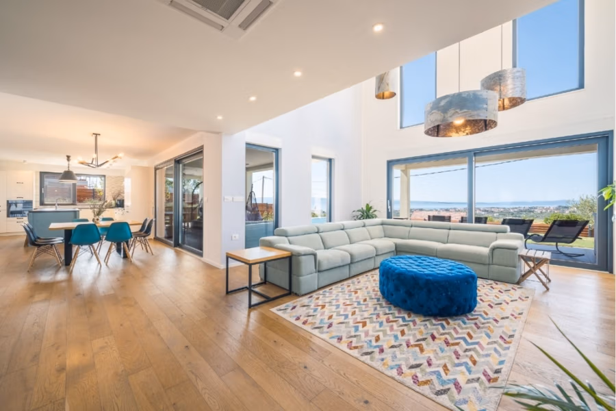 Villa am Stadtrand von Split großzügige Aufteilung, hohe Räume und beheiztem Pool - Geräumiges Wohnzimmer mit hohen Decken, großen Fenstern und Holzboden. Hellgraues Ecksofa, blauer Polsterhocker und farbenfroher Teppich. Offener Essbereich mit türkisfarbenen Stühlen. Blick auf die Küste durch Glastüren. Moderne Einrichtung. (41931)