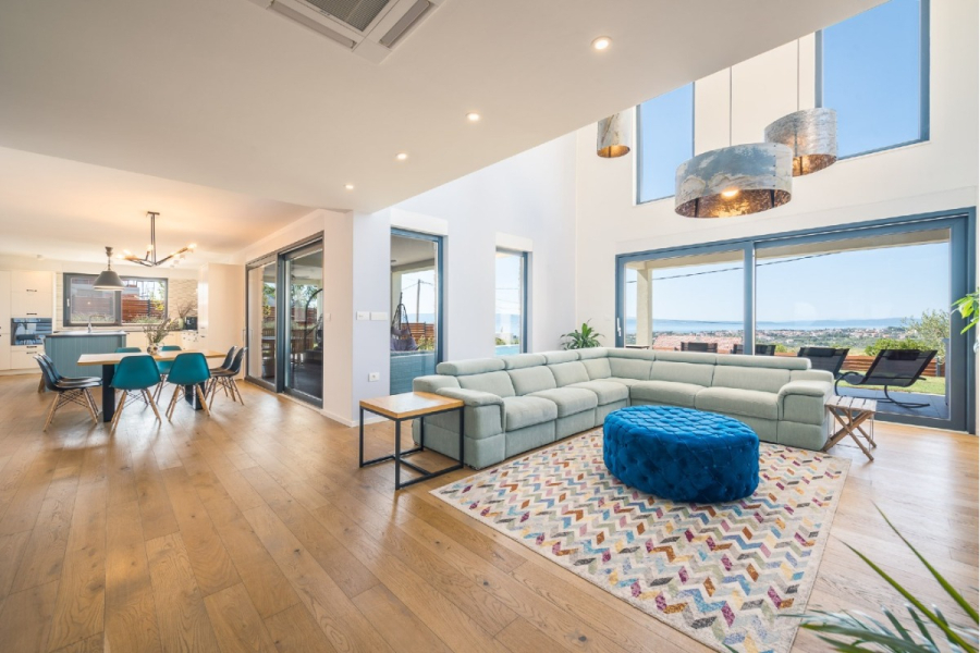 Villa am Stadtrand von Split großzügige Aufteilung, hohe Räume und beheiztem Pool - Moderner offener Wohn- und Essbereich mit großen Fenstern, hellem Holzfußboden, einem hellen Sektionssofa, einer blauen Ottomane, einem farbenfrohen Teppich und einem Esstisch mit Stühlen in Petrol. Durch die Fenster kann man das Meer sehen. (46560)