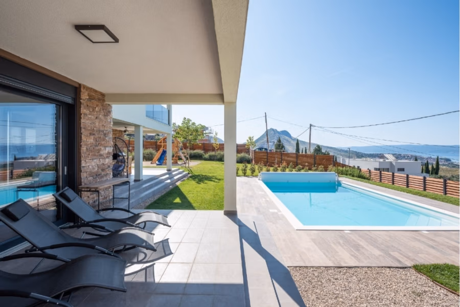 Villa am Stadtrand von Split großzügige Aufteilung, hohe Räume und beheiztem Pool - Außenterrasse mit schwarzen Liegen neben einem klaren, blauen Swimmingpool. Im Garten steht ein Holzspielgerät. Im Hintergrund sind Berge und blauer Himmel zu sehen. Modernes Haus mit Stein- und Glaselementen. (41928)
