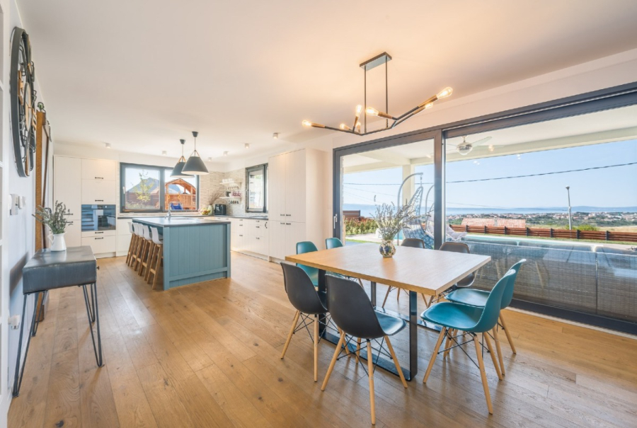 Villa am Stadtrand von Split großzügige Aufteilung, hohe Räume und beheiztem Pool - Moderne, offene Küche und Essbereich mit Holzfußboden, blauer Insel, zeitgenössischen Leuchten und großen Glastüren, die zu einer Terrasse mit Sitzgelegenheiten im Freien und einer malerischen Aussicht führen. (46552)