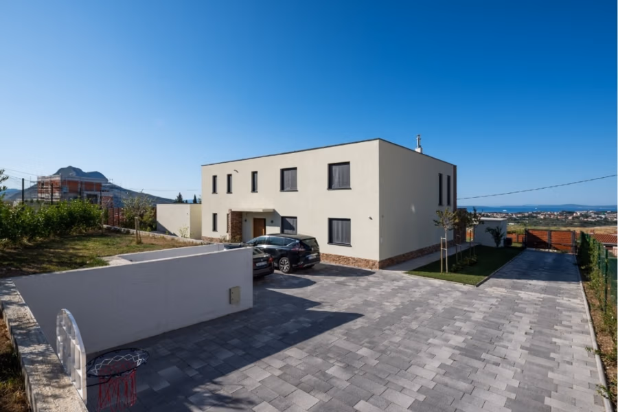 Villa am Stadtrand von Split großzügige Aufteilung, hohe Räume und beheiztem Pool - Modernes zweistöckiges Haus mit Flachdach, hellbeiger Fassade und dunklen Zierelementen. Ein Auto parkt in der gepflasterten Einfahrt. Das Grundstück ist eingezäunt und hat einen kleinen Garten an der Seite. Im Hintergrund sind Berge und eine Stadt unter einem klaren blauen Himmel zu sehen. (41926)