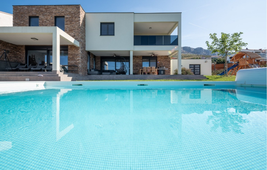 Villa am Stadtrand von Split großzügige Aufteilung, hohe Räume und beheiztem Pool - Modernes zweistöckiges Haus mit einer großen Terrasse, einem Essbereich im Freien und einem sauberen Swimmingpool im Vordergund. Der Hinterhof hat einen grünen Rasen und einen Baum, mit Bergen im Hintergrund sichtbar. (46549)