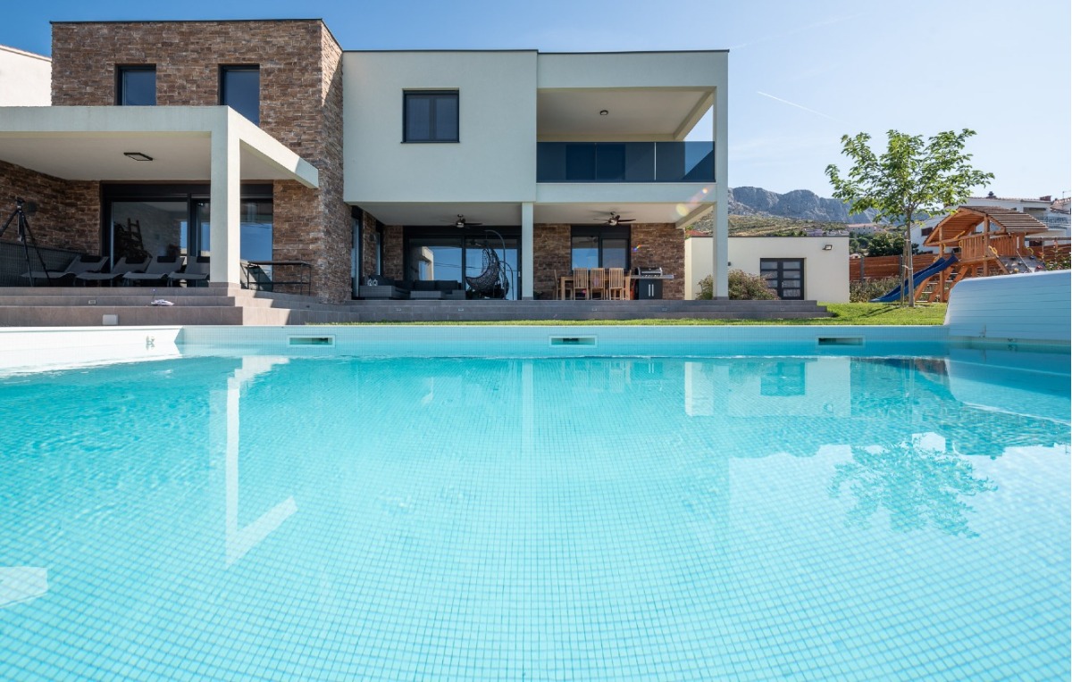Modernes zweistöckiges Haus mit einer großen Terrasse, einem Essbereich im Freien und einem sauberen Swimmingpool im Vordergund. Der Hinterhof hat einen grünen Rasen und einen Baum, mit Bergen im Hintergrund sichtbar. - Villa am Stadtrand von Split großzügige Aufteilung, hohe Räume und beheiztem Pool
