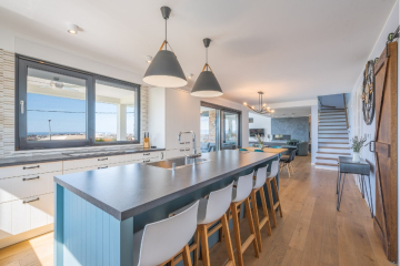 Helle, moderne Küche mit einer langen blauen Insel, weißen Stühlen, Pendelleuchten, hellen Holzböden, großen Fenstern und einem offenen Grundriss, der zu einem Wohnbereich und einer Treppe führt. - Villa am Stadtrand von Split großzügige Aufteilung, hohe Räume und beheiztem Pool