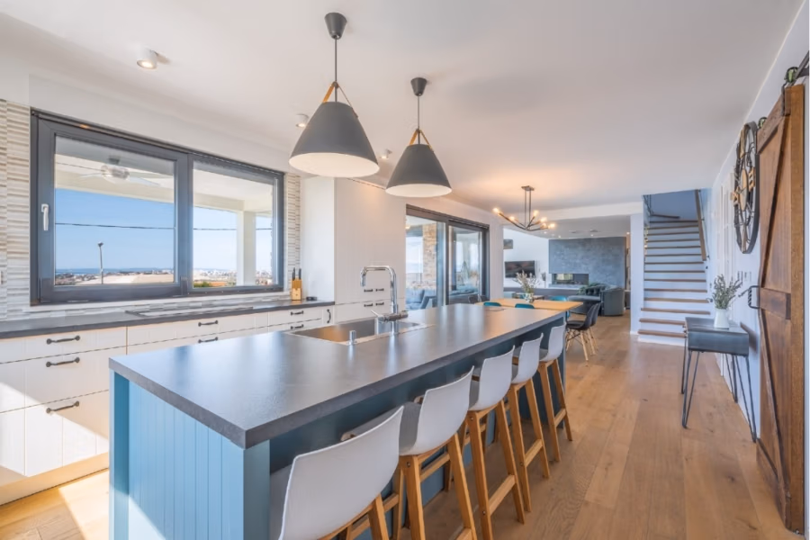 Villa am Stadtrand von Split großzügige Aufteilung, hohe Räume und beheiztem Pool - Geräumige, moderne Küche mit langer, blauer Kücheninsel und weißen Hockern. Darüber hängen Pendelleuchten. Große Fenster sorgen für natürliches Licht. Holzböden, weiße Schränke und ein offener Essbereich im Hintergrund mit einer Treppe nach oben. (41923)