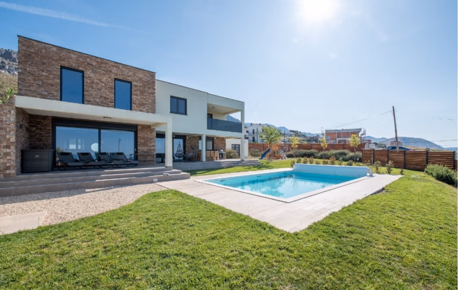 Villa am Stadtrand von Split großzügige Aufteilung, hohe Räume und beheiztem Pool - Modernes zweistöckiges Haus mit einer Mischung aus Stein und weißer Fassade und großen Glasfenstern. Im Vordergrund ein Swimmingpool und eine grüne Wiese unter einem strahlend blauen Himmel mit strahlender Sonne. Im Hintergrund sind Hügel und andere Häuser zu sehen. (41927)
