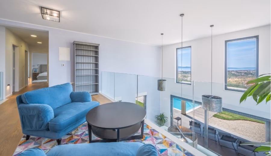 Villa am Stadtrand von Split großzügige Aufteilung, hohe Räume und beheiztem Pool - Modernes Wohnzimmer mit blauen Samtsitzen und einem runden schwarzen Couchtisch auf einem farbenfrohen geometrischen Teppich. Große Fenster bieten Blick auf einen Swimmingpool und die Landschaft. Moderne Hängelampen und ein graues Bücherregal runden den Raum ab. (41932)