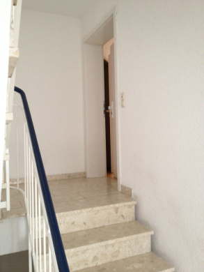 Eine Treppe mit beigen Marmorstufen und einem blauen Handlauf führt zu einem weißen Flur mit einer offenen weißen Tür am oberen Ende. Die Wände und die Umgebung sind schlicht und minimalistisch. - Provisionsfrei: 3,5 Zimmer Wohnung 90m² / 150m vom Bodensee entfernt mit Garten, Garage, …