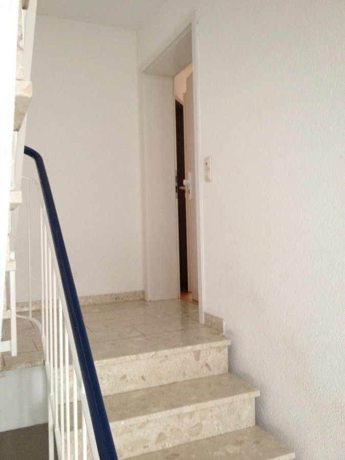 Provisionsfrei: 3,5 Zimmer Wohnung 90m² / 150m vom Bodensee entfernt mit Garten, Garage, … - Eine Treppe mit beigen Marmorstufen und einem blauen Handlauf führt zu einem weißen Flur mit einer offenen weißen Tür am oberen Ende. Die Wände und die Umgebung sind schlicht und minimalistisch. (45567)