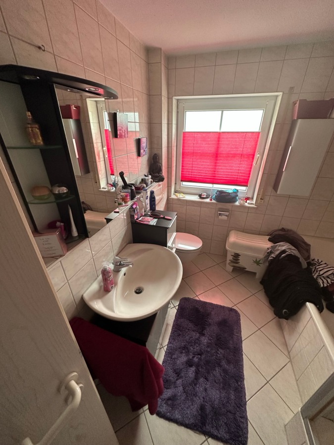 Provisionsfrei: 3,5 Zimmer Wohnung 90m² / 150m vom Bodensee entfernt mit Garten, Garage, … - Ein kleines, gefliestes Badezimmer mit Waschbecken, Spiegel, Toilette und einem Fenster, das mit einer roten Jalousie abgedeckt ist. Auf dem Waschbecken und den Regalen befinden sich verschiedene Toilettenartikel. Ein dunkelvioletter Teppich und ein Handtuch liegen auf dem Boden, und auf einer Bank liegt Wäsche. (45575)