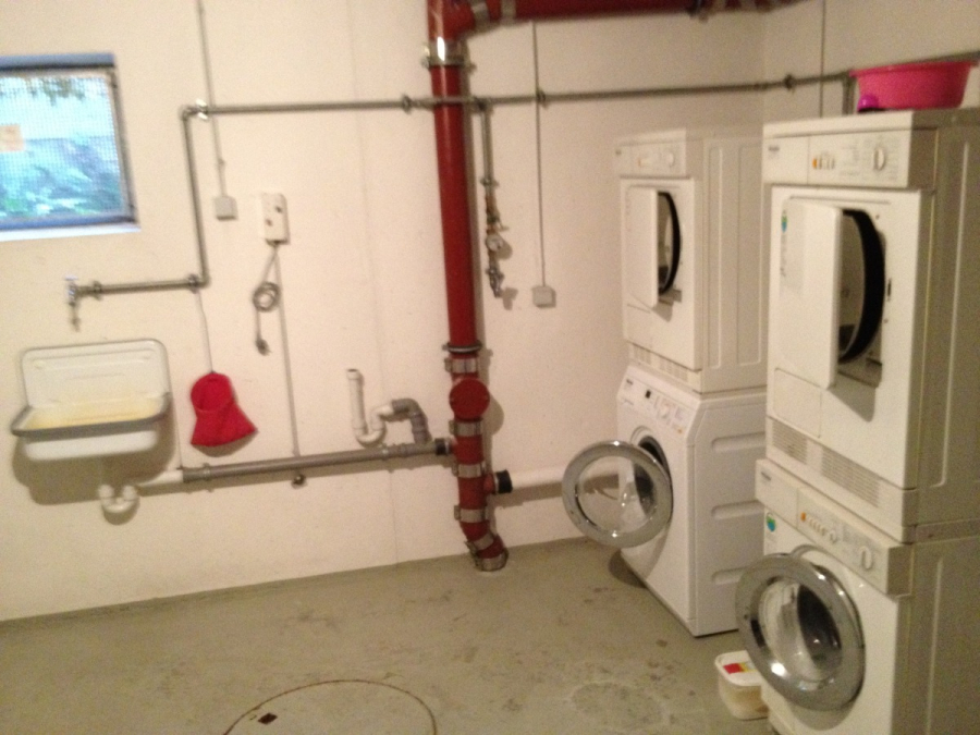 Provisionsfrei: 3,5 Zimmer Wohnung 90m² / 150m vom Bodensee entfernt mit Garten, Garage, … - Eine Waschküche mit zwei gestapelten Waschmaschinen und Trocknern, zwei offenen Waschmaschinentüren, einem Waschbecken, freiliegenden Rohren, einem roten Feuerlöschrohr und einem Eimer auf dem Boden. An der linken Wand befindet sich ein kleines Fenster. (45581)