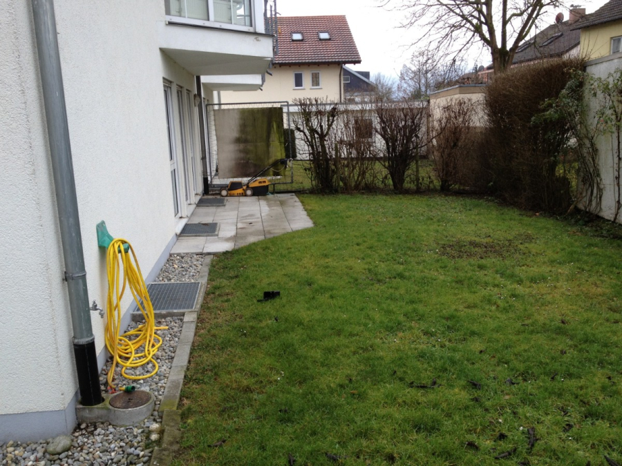 Provisionsfrei: 3,5 Zimmer Wohnung 90m² / 150m vom Bodensee entfernt mit Garten, Garage, … - Ein kleiner Hinterhof mit grünem Gras, einem gelben Gartenschlauch auf einem Aufroller an der Mauer, einer gepflasterten Terrasse und einem Rasenmäher auf der Terrasse. Sträucher und ein Baum säumen den Zaun auf der Rückseite. (45577)