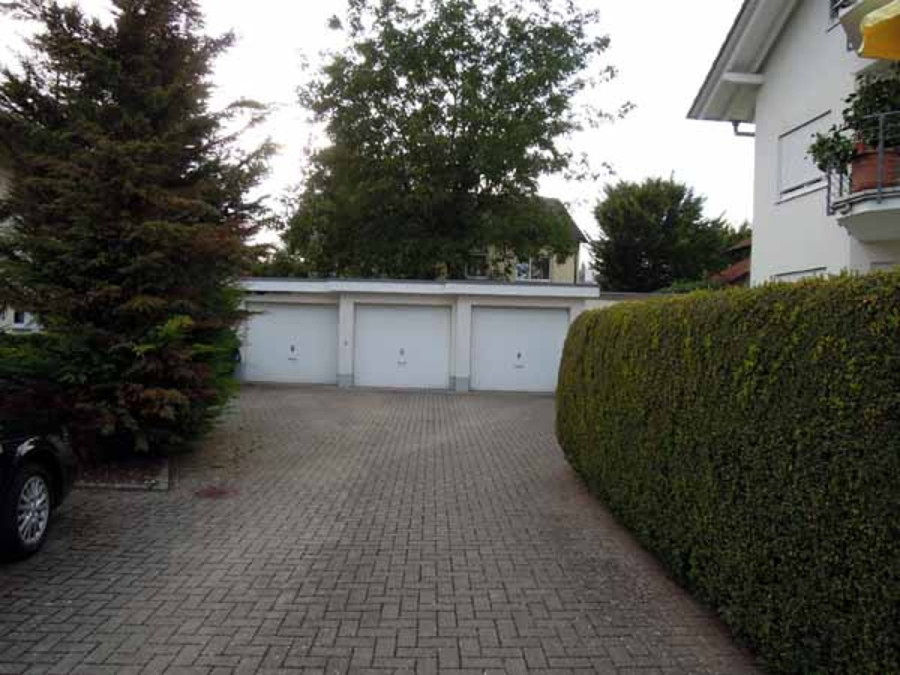 Provisionsfrei: 3,5 Zimmer Wohnung 90m² / 150m vom Bodensee entfernt mit Garten, Garage, … - Eine gepflasterte Einfahrt führt zu drei weißen Garagentoren in einer Reihe, die von einer hohen grünen Hecke auf der rechten Seite und Bäumen im Hintergrund begrenzt werden. Ein weißes Wohngebäude und ein geparktes Auto sind teilweise an den Seiten zu sehen. (45582)