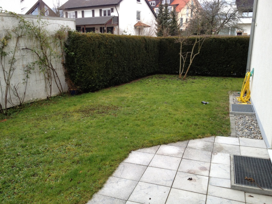 Provisionsfrei: 3,5 Zimmer Wohnung 90m² / 150m vom Bodensee entfernt mit Garten, Garage, … - Ein kleiner Hinterhof mit einem grasbewachsenen Rasen, einer gefliesten Terrasse und einem gelben Gartenschlauch auf der rechten Seite. Hecken säumen die Rückseite und die Seite, im Hintergrund sind ein paar kahle Sträucher und die Nachbarhäuser zu sehen. (45576)
