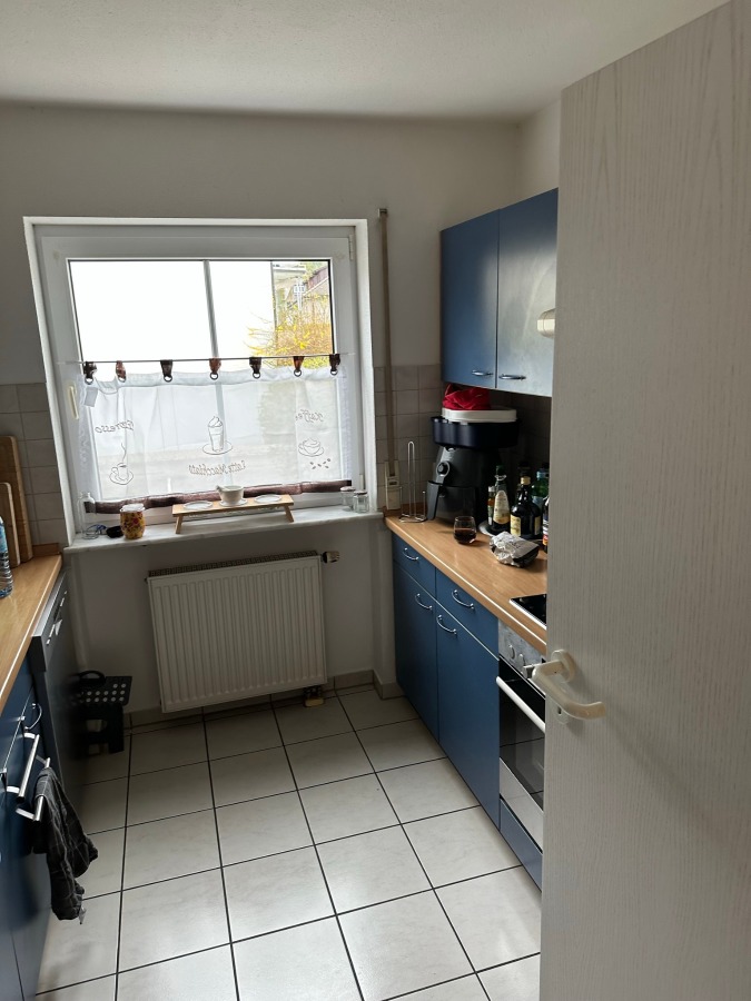 Provisionsfrei: 3,5 Zimmer Wohnung 90m² / 150m vom Bodensee entfernt mit Garten, Garage, … - Kleine Küche mit blauen Schränken, hölzernen Arbeitsflächen, einem Fenster mit durchsichtigem Vorhang, verschiedenen Küchenutensilien auf den Arbeitsflächen, einer Kaffeemaschine und Flaschen neben dem Spülbecken. Der Boden ist gefliest, und eine weiße Tür ist teilweise geöffnet. (45572)
