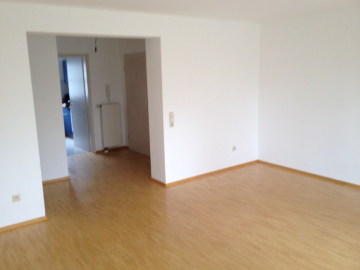 Ein leerer Raum mit hellem Holzboden, weißen Wänden und einer breiten Tür, die zu einem Flur und einem weiteren Raum führt. Der Raum ist unmöbliert und hat ein sauberes, minimalistisches Aussehen. - Provisionsfrei: 3,5 Zimmer Wohnung 90m² / 150m vom Bodensee entfernt mit Garten, Garage, …