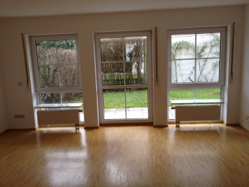 Ein heller, leerer Raum mit hellem Holzfußboden, drei großen Fenstern und zwei Heizkörpern. Die Fenster bieten einen Blick auf einen grasbewachsenen Außenbereich mit Sträuchern und einer weißen Wand. - Provisionsfrei: 3,5 Zimmer Wohnung 90m² / 150m vom Bodensee entfernt mit Garten, Garage, …