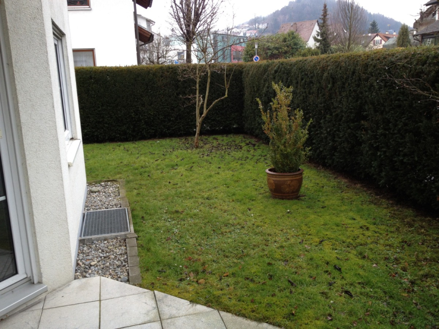 Provisionsfrei: 3,5 Zimmer Wohnung 90m² / 150m vom Bodensee entfernt mit Garten, Garage, … - Ein kleiner, eingezäunter Garten mit grünem Gras, einem Topfstrauch und einem blattlosen Baum, umrahmt von hohen Hecken und einem weißen Gebäude, mit Häusern und Hügeln im Hintergrund. (45580)