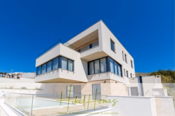 Modernes, weißes, dreistöckiges Haus mit großen Fenstern und Außenterrassen unter strahlend blauem Himmel. Ein Glaszaun umgibt einen Terrassenbereich im Vordergrund. Unten ist das Logo von „Hike Homes“ mit „TRAUM-FERIENIMMOBILIEN IN KROATIEN“ zu sehen. - Ein Paradies mit atemberaubendem Meerblick! 4 SZ, Pool, Solar, …