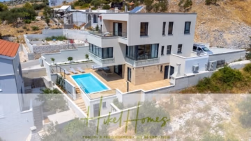 Eine moderne weiße Villa mit Terrasse und Swimmingpool liegt an einem Hang, umgeben von spärlicher Vegetation und anderen Häusern. Die Villa zeichnet sich durch große Fenster und ein mehrstöckiges Design aus. Unten ist ein Logo mit dem Text „Mike Homes“ eingeblendet. - Ein Paradies mit atemberaubendem Meerblick! 4 SZ, Pool, Solar, …