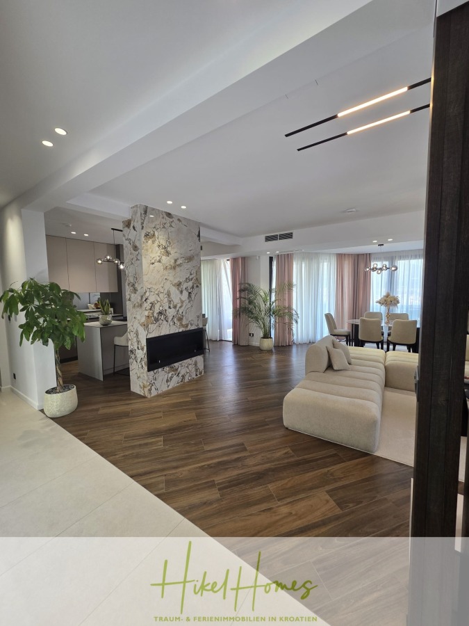 Luxus trifft Meerblick – Ein Anwesen der Extraklasse – Sofort einziehen - Ein moderner, offener Wohn- und Essbereich mit Holzfußboden, einem Marmorkamin, großen Fenstern mit transparenten Vorhängen, neutraler Einrichtung und Zimmerpflanzen. Das "Hiked Homes"-Logo ist am Boden angebracht. (46604)