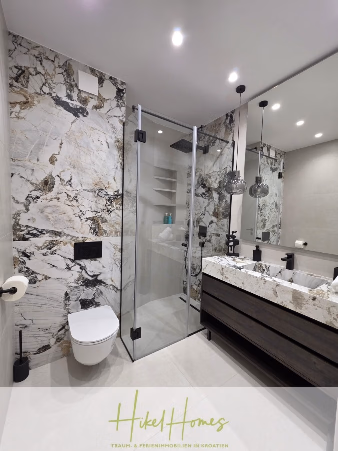 Luxus trifft Meerblick – Ein Anwesen der Extraklasse – Sofort einziehen - Modernes Badezimmer mit Marmorwand und Glasduschkabine. Weiße Toilette neben der Dusche, dunkler Holzwaschtisch mit Marmorplatte, runde Waschbecken und Hängelampen. Minimalistisches Design in neutralen Tönen. (43708)