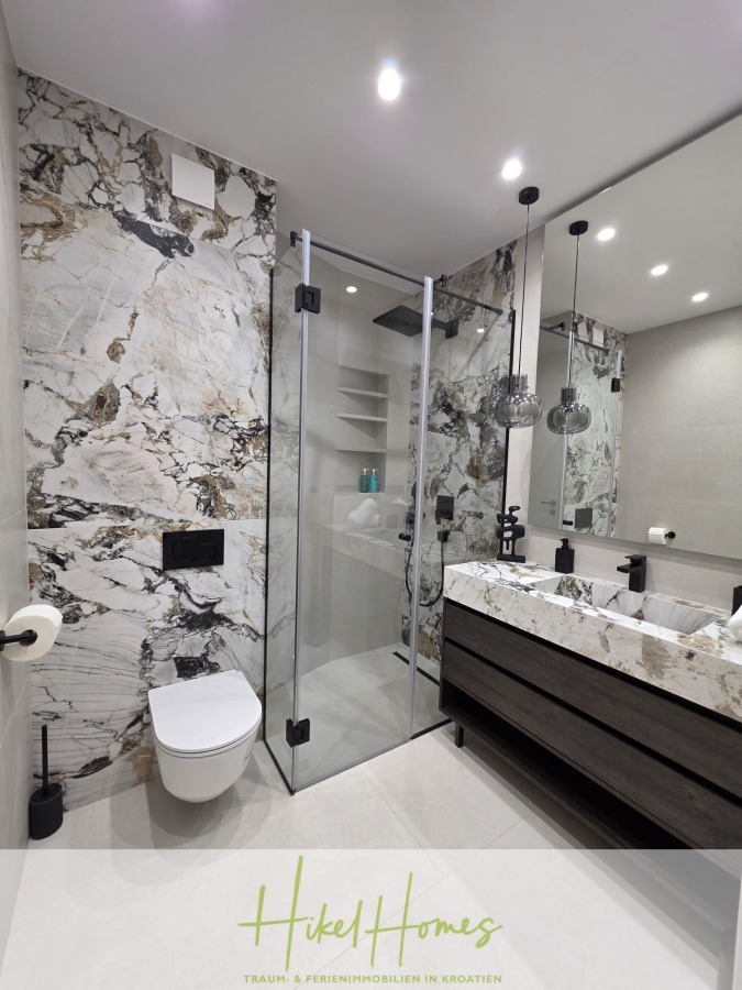 Luxus trifft Meerblick – Ein Anwesen der Extraklasse – Sofort einziehen - Modernes Badezimmer mit Marmorwänden, einer Glasduschkabine, einer wandmontierten Toilette, einem schwimmenden Waschbecken, einem großen Spiegel und Pendelleuchten. Der Raum ist hell und zeitgemäß mit schlanken Armaturen. (46616)