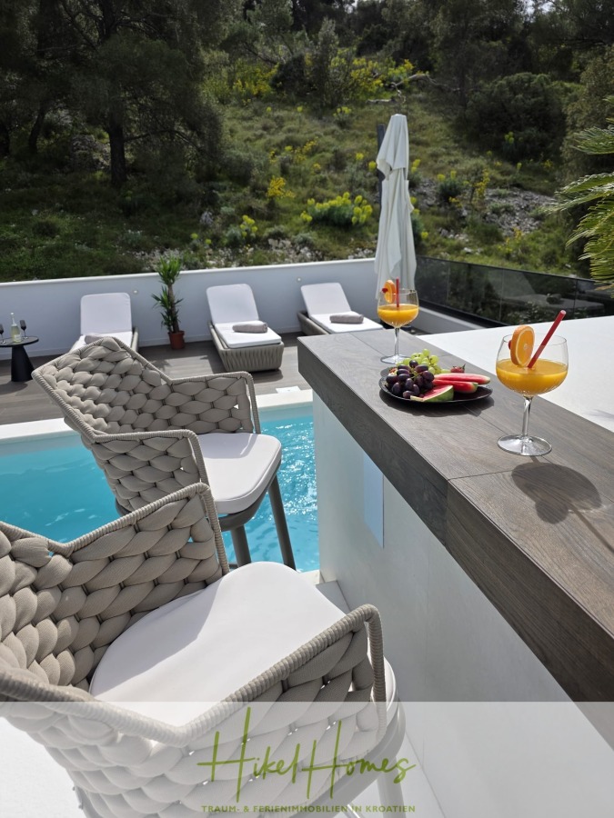 Luxus trifft Meerblick – Ein Anwesen der Extraklasse – Sofort einziehen - Eine luxuriöse Poolszene mit geflochtenen Stühlen an einer Bar, zwei Orangencocktails mit Fruchtbeilagen und einem Teller mit frischem Obst. Im Hintergrund sind Liegestühle und Bäume zu sehen. (46619)