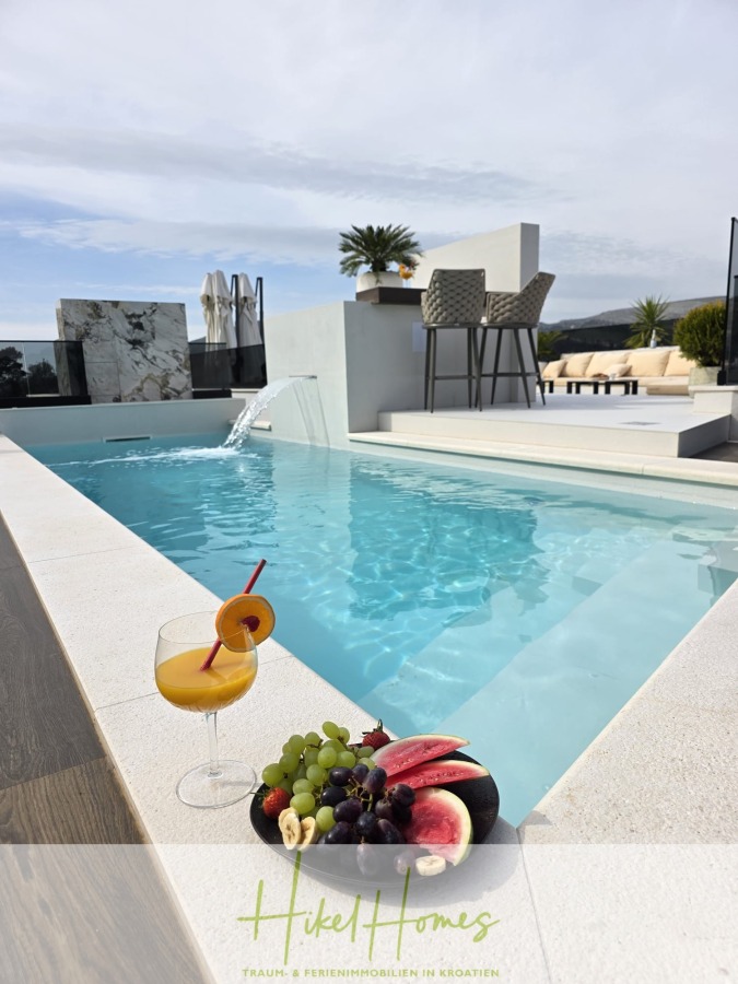 Luxus trifft Meerblick – Ein Anwesen der Extraklasse – Sofort einziehen - Eine erfrischende Poolszene mit einem modernen Swimmingpool, einem Getränk mit einer Orangenscheibe und einer Kirsche sowie einem Teller mit Obst am Rand. Im Hintergrund sind Liegestühle, Barhocker und Palmen unter einem bewölkten Himmel zu sehen. (46606)