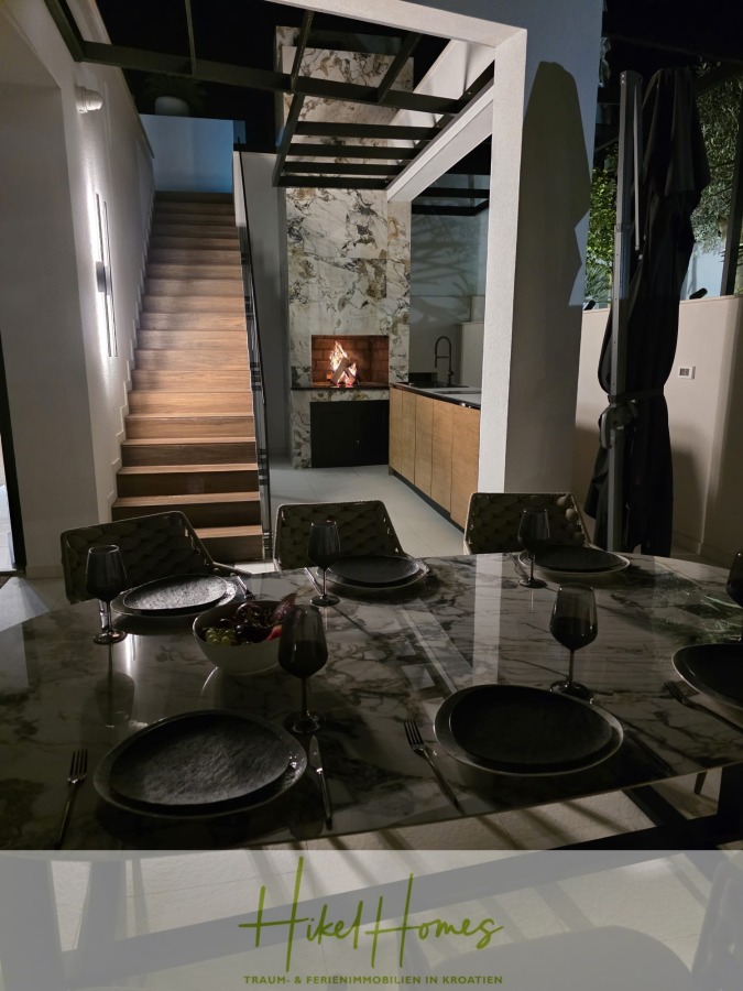 Luxus trifft Meerblick – Ein Anwesen der Extraklasse – Sofort einziehen - Ein moderner nächtlicher Essbereich zeigt einen Marmortisch mit schwarzem Geschirr und Gläsern, eine Treppe mit beleuchteten Stufen und eine Küche mit einem beleuchteten Kamin im Hintergrund. Der Raum ist schlicht und modern eingerichtet. (46614)