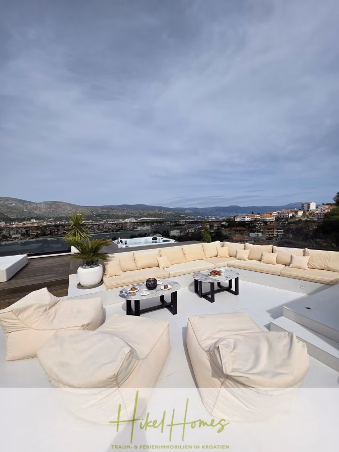 Luxus trifft Meerblick – Ein Anwesen der Extraklasse – Sofort einziehen - Eine Dachterrasse mit einem geschwungenen, beigefarbenen Sofa und passenden Poufs. Schwarze Couchtische mit Snacks stehen auf einer weißen Plattform. Unter einem bewölkten Himmel bietet sich ein Panoramablick auf eine Stadt und Hügel. Auf der linken Seite steht eine Topfpflanze. (43691)