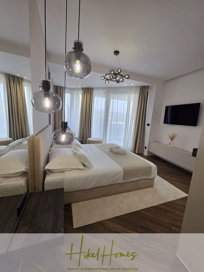 Luxus trifft Meerblick – Ein Anwesen der Extraklasse – Sofort einziehen - Modernes Schlafzimmer mit großem Bett, Hängelampen und Wandfernseher. Das bodentiefe Fenster ist mit transparenten Vorhängen und einem schicken Kronleuchter ausgestattet. Das Zimmer ist in neutralen Farbtönen gehalten und hat einen glänzenden Holzboden. (43700)