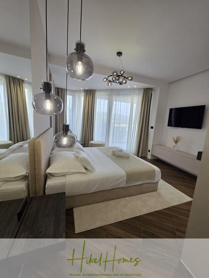 Luxus trifft Meerblick – Ein Anwesen der Extraklasse – Sofort einziehen - Modernes Schlafzimmer mit großen Fenstern und durchsichtigen Vorhängen, neutralen Farbtönen, einem Doppelbett mit weißer Bettwäsche, hängenden Glaspendelleuchten, einem an der Wand montierten Fernseher und zeitgenössischem Dekor. Das Logo "Hikel Homes" erscheint am unteren Rand. (46607)
