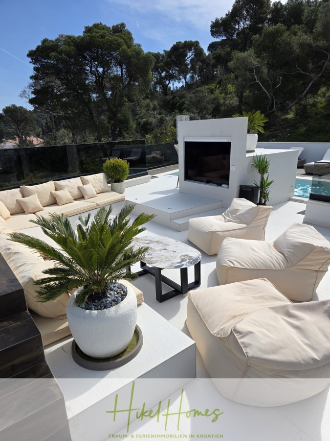 Luxus trifft Meerblick – Ein Anwesen der Extraklasse – Sofort einziehen - Moderne Außenterrasse mit cremefarbenen Sofas, kleinen Tischen, einer Topfpalme und einem großen Flachbildfernseher. Umgeben von üppigen Bäumen ist der Raum hell und stilvoll, ideal zum Entspannen oder für Gäste. Logo "Hiket Homes" am unteren Rand. (46599)