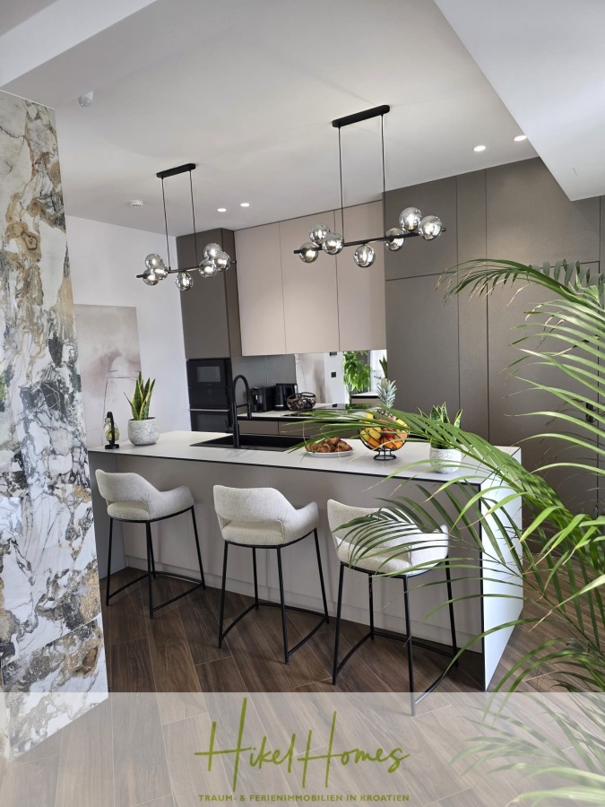 Luxus trifft Meerblick – Ein Anwesen der Extraklasse – Sofort einziehen - Moderne Küche mit weißer Kochinsel, drei gepolsterten Barhockern, Metallleuchtern, grauen Schränken und dekorativen Pflanzen. Eine Marmorwand und eine Obstschale bringen Farbe und Textur in das schlichte, moderne Design. (46611)