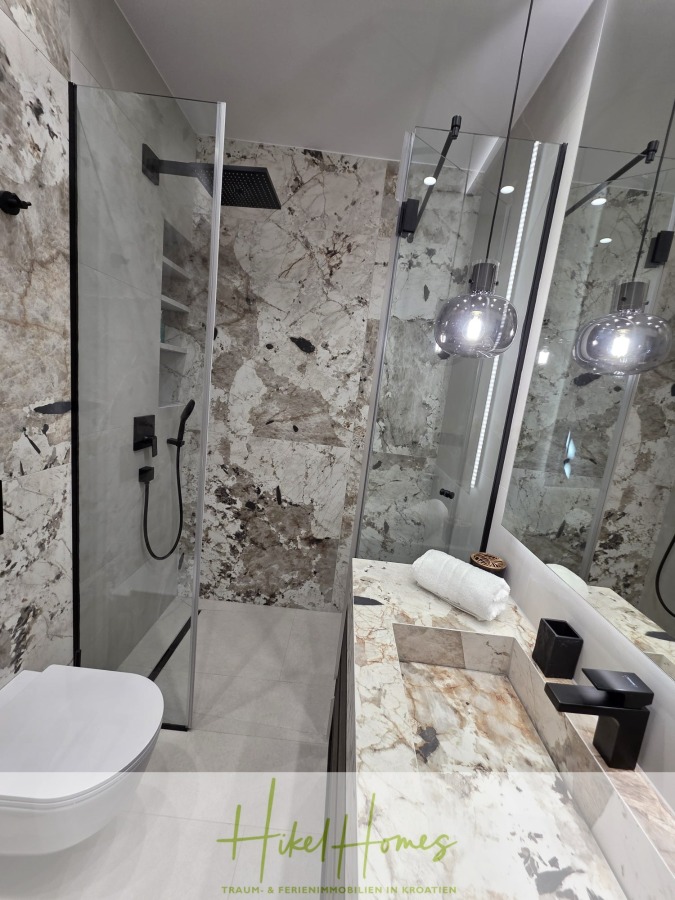 Luxus trifft Meerblick – Ein Anwesen der Extraklasse – Sofort einziehen - Modernes Badezimmer mit Marmorwänden und -arbeitsplatte, Glasduschabtrennung mit schwarzen Armaturen, Pendelleuchten, weißer Toilette und weißen Handtuchrollen auf dem Waschbecken. Die Spiegelung zeigt ein schlichtes und elegantes Design. (46618)