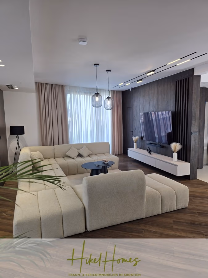 Luxus trifft Meerblick – Ein Anwesen der Extraklasse – Sofort einziehen - Modernes Wohnzimmer mit einem großen cremefarbenen Sofa, einem runden schwarzen Couchtisch und einem Flachbildfernseher an einer getäfelten Akzentwand. Eine Seite ist mit raumhohen Vorhängen bedeckt, und Pendelleuchten hängen von der Decke. Brauner Holzboden. (43696)