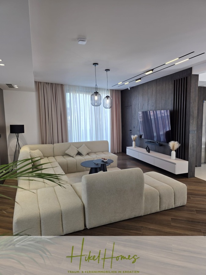 Luxus trifft Meerblick – Ein Anwesen der Extraklasse – Sofort einziehen - Ein modernes Wohnzimmer mit einem großen beigen L-förmigen Sofa, einem runden Couchtisch, einem Flachbildfernseher an einer dunklen Akzentwand und raumhohen Vorhängen, die den Raum mit natürlichem Licht erhellen. (46603)