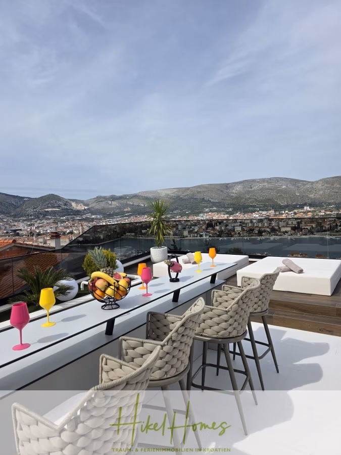 Luxus trifft Meerblick – Ein Anwesen der Extraklasse – Sofort einziehen - Dachterrasse mit einem langen Esstisch, gedeckt mit bunten Gläsern und Obstschalen. Um den Tisch herum stehen bequeme, geflochtene Barhocker. Im Hintergrund sind ein malerischer Blick auf die Berge und Gebäude unter einem bewölkten Himmel zu sehen. (43694)