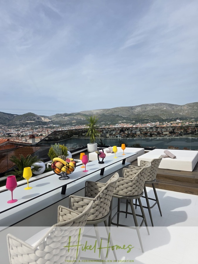 Luxus trifft Meerblick – Ein Anwesen der Extraklasse – Sofort einziehen - Eine moderne Dachterrasse mit geflochtenen Stühlen, einem langen Esstisch mit bunten Gläsern, Obst und Pflanzen, mit Blick auf die Stadt und die Berge unter einem teilweise bewölkten Himmel. (46600)