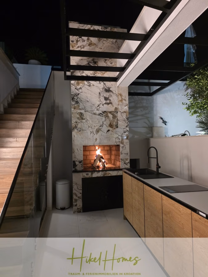 Luxus trifft Meerblick – Ein Anwesen der Extraklasse – Sofort einziehen - Moderner Innenraum mit einem Kamin aus Marmor und brennendem Feuer neben einer Küchentheke aus Holz mit Spüle. Treppen mit Holzstufen und Pflanzen sorgen für eine gemütliche Atmosphäre. (43705)