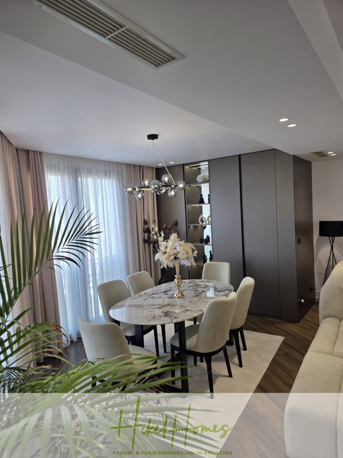 Luxus trifft Meerblick – Ein Anwesen der Extraklasse – Sofort einziehen - Ein moderner Essbereich mit einem runden Marmortisch, sechs beigefarbenen Polsterstühlen, einem dekorativen Vasenaufsatz, einer modernen Hängelampe, bodentiefen Fenstern mit Vorhängen und einem Dekor in neutralen Tönen. (46610)