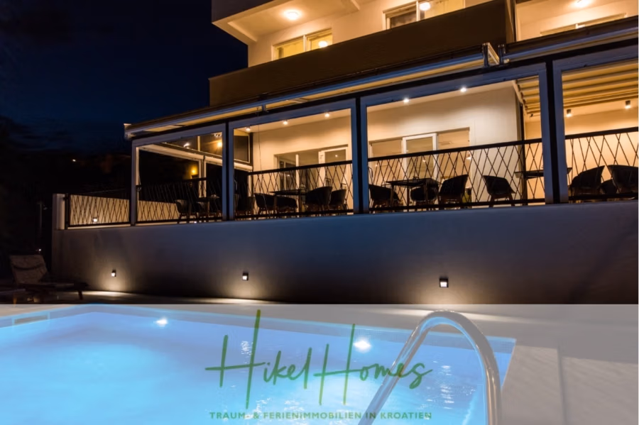 340m² Villa mit Pool auf der Insel Ciovo – vollständig möbiliert – auch Boutiquehotel möglich - Ein modernes Gebäude mit einem beleuchteten Pool im Vordergrund. Das Gebäude verfügt über einen Balkon mit Stühlen und eine Reihe von Fenstern. Das Logo unten lautet „Hikel Homes“. Die Szene suggeriert eine entspannte Urlaubsatmosphäre, möglicherweise in Kroatien. (41644)
