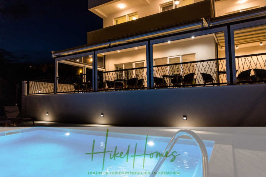 340m² Villa mit Pool auf der Insel Ciovo – vollständig möbiliert – auch Boutiquehotel möglich - Eine moderne, gut beleuchtete Villa mit einem geräumigen Balkon blickt auf ein leuchtendes Außenschwimmbad bei Nacht. Das Logo "Hikel Homes" und der Text "TRAUM FERIENIMMOBILIEN IN KROATIEN" erscheinen am unteren Rand. (45557)