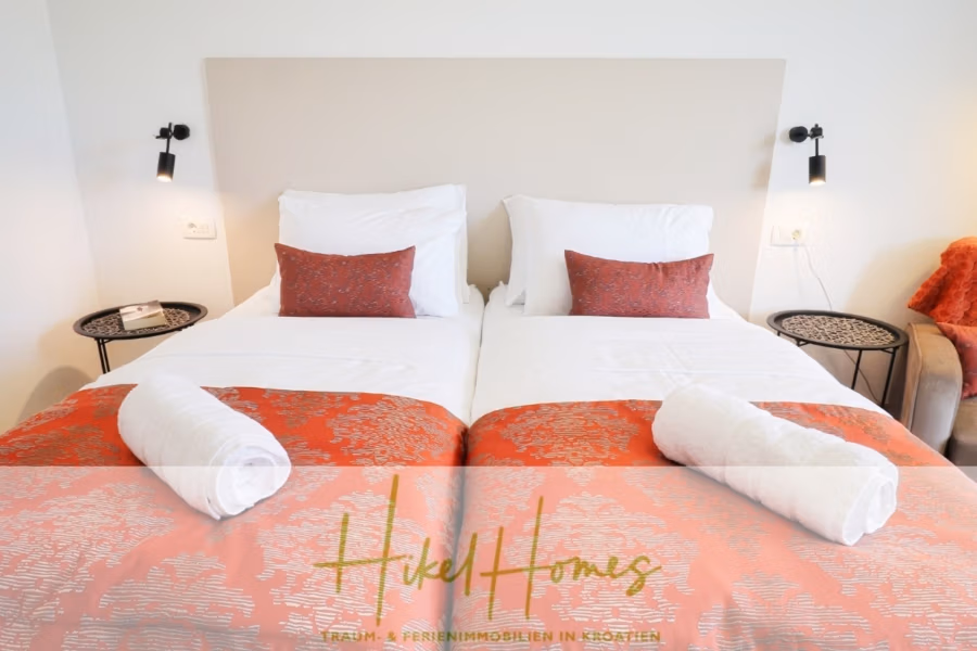 340m² Villa mit Pool auf der Insel Ciovo – vollständig möbiliert – auch Boutiquehotel möglich - Zwei Einzelbetten mit weißer Bettwäsche und orangefarbenen Läufern stehen nebeneinander. Jedes Bett verfügt über ein orangefarbenes Kissen und ein gerolltes weißes Handtuch. Schwarze Wandleuchten sorgen über runden Beistelltischen für Licht. Unten steht „Hotel Homes“. (41636)