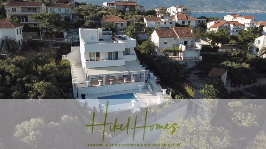 340m² Villa mit Pool auf der Insel Ciovo – vollständig möbiliert – auch Boutiquehotel möglich - Luftaufnahme eines Hangviertels mit weißen Häusern und roten Dächern. Ein modernes Haus mit Pool davor. Grüne Bäume umgeben das Gebiet, in der Ferne ist eine Bergkette zu sehen. Der Text lautet „Hikel Homes“ in grüner Schreibschrift. (41634)