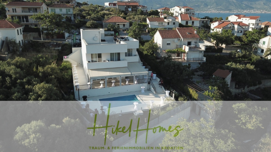 340m² Villa mit Pool auf der Insel Ciovo – vollständig möbiliert – auch Boutiquehotel möglich - Eine moderne weiße Villa mit Pool ist umgeben von Häusern im mediterranen Stil und Grünanlagen auf einem Hügel mit Blick auf das Meer in Kroatien. Das Bild zeigt das Logo und den Text: "Hikel Homes - Traum- & Ferienimmobilien in Kroatien. (45544)