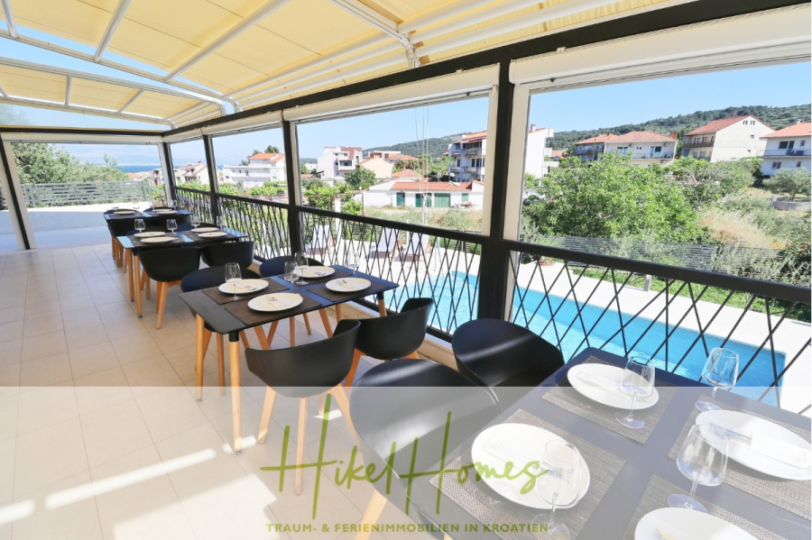 340m² Villa mit Pool auf der Insel Ciovo – vollständig möbiliert – auch Boutiquehotel möglich - Ein überdachter Essbereich im Freien mit schwarzen Stühlen und Tischen für eine Mahlzeit mit Blick auf einen Swimmingpool und eine Nachbarschaft von Häusern mit Grün und Hügeln im Hintergrund. Der Text lautet "Hike Homes" auf Deutsch. (45532)
