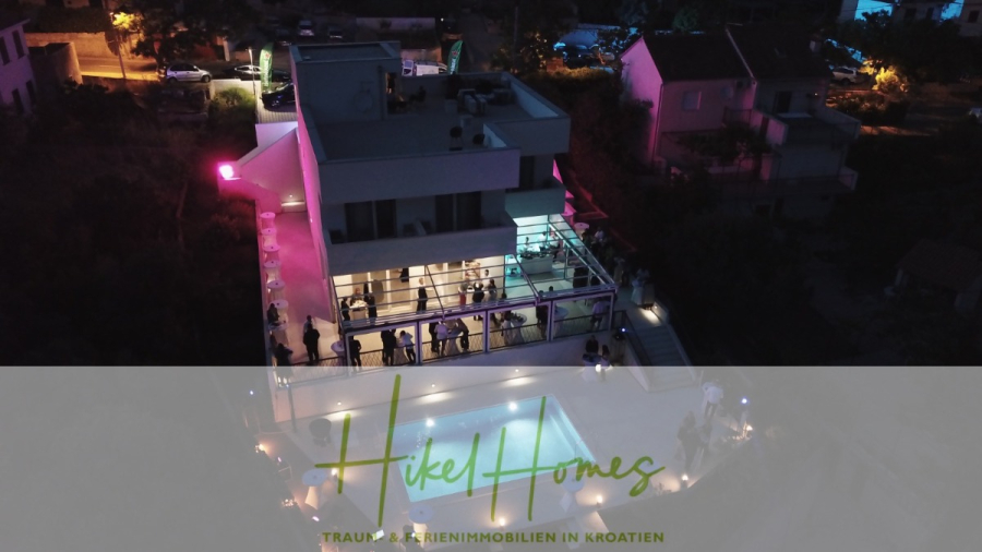 340m² Villa mit Pool auf der Insel Ciovo – vollständig möbiliert – auch Boutiquehotel möglich - Ein modernes, mehrstöckiges Haus in Kroatien ist nachts mit bunten Lichtern beleuchtet. Die Menschen sind auf dem Balkon und um den Pool versammelt. Das Logo "Hikel Homes" und deutscher Text erscheinen am unteren Rand. (45552)